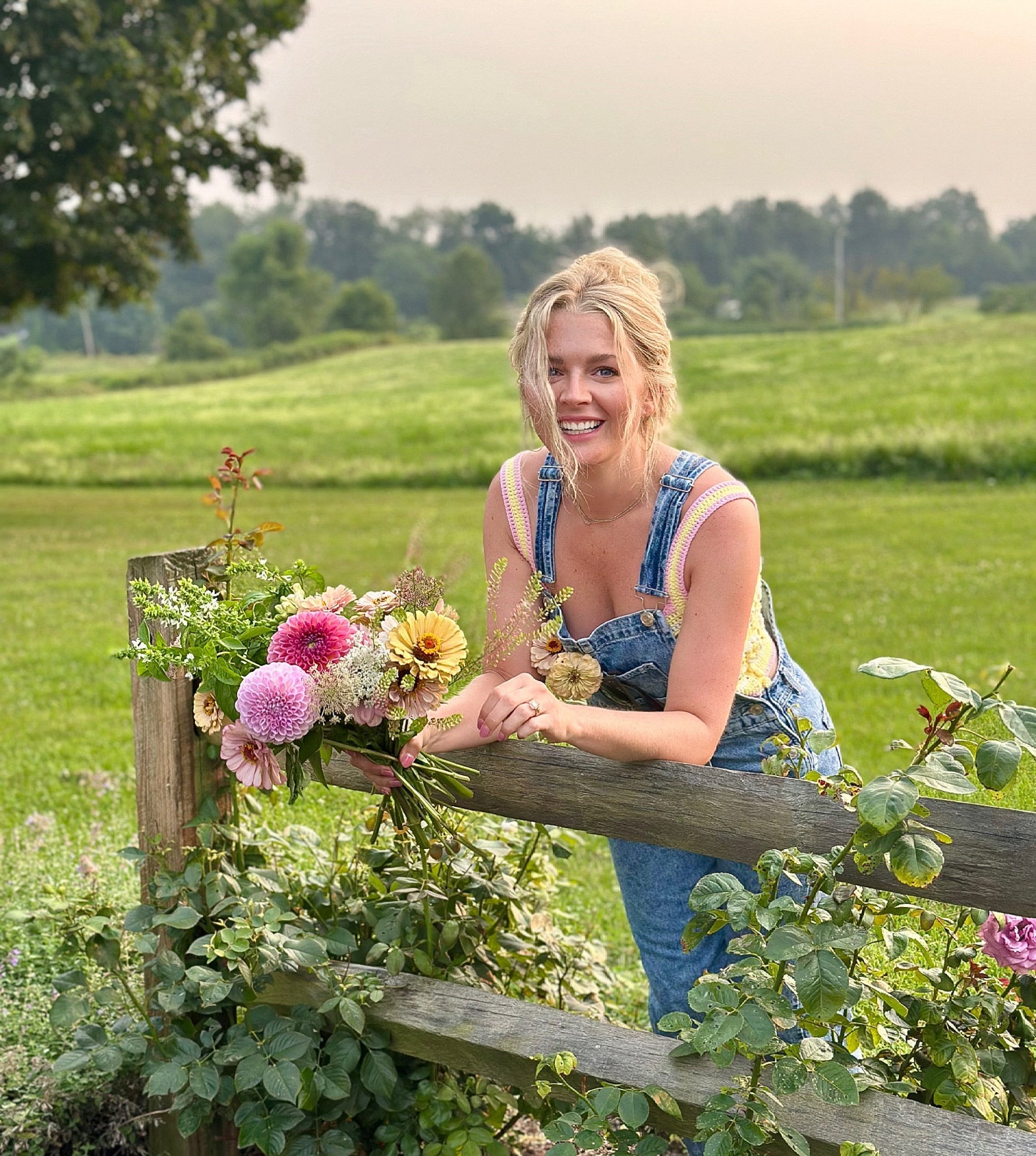 Woman in overalls leaning on a wooden fence with flowers in a field Solera Flower Farm